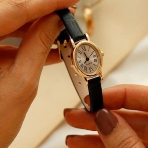 May include: A wristwatch with an oval gold-tone case and a white dial featuring black Roman numerals. The watch has a thin black textured leather strap and a quartz movement. The word QUARTZ is printed in black on the center of the watch face.