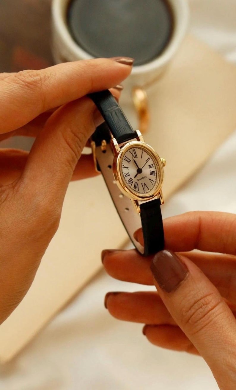 May include: A wristwatch with an oval gold-tone case and a white dial featuring black Roman numerals. The watch has a thin black textured leather strap and a quartz movement. The word QUARTZ is printed in black on the center of the watch face.