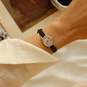 May include: A gold-toned oval wristwatch featuring a white dial with black Roman numeral hour markers and thin black hands. The watch is secured with a slim, smooth black leather strap that wraps around the wrist, providing a classic look.