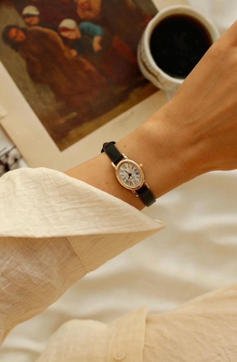 May include: A gold-toned oval wristwatch featuring a white dial with black Roman numeral hour markers and thin black hands. The watch is secured with a slim, smooth black leather strap that wraps around the wrist, providing a classic look.