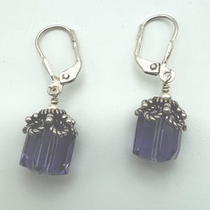 May include: A pair of drop earrings featuring faceted, deep purple, cube-shaped crystals. Each crystal is topped with a decorative, oxidized silver-toned metal cap with a twisted rope texture and small bead accents, suspended from silver lever-back hooks.