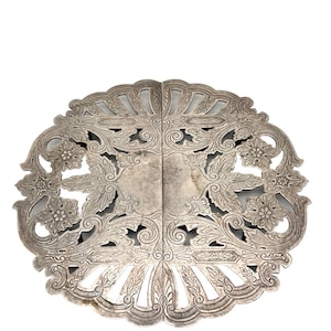 May include: A circular silver-toned metal trivet featuring an intricate openwork design with embossed floral motifs, scrolling leaf patterns, and a scalloped edge. The piece is composed of two symmetrical halves joined together.