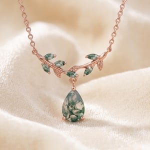 May include: A rose gold necklace featuring a delicate branch-like design adorned with small, green moss agate leaf-shaped accents. A single, pear-shaped moss agate gemstone hangs as a central pendant from the center of the branch structure.