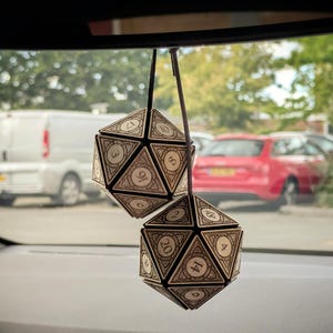 May include: Two hanging 20-sided dice ornaments suspended from a dark cord. Each die is constructed from light-coloured wood panels with dark edges and intricate engraved patterns. The faces feature clearly visible black numbers including 3, 8, 16, 17, 2, 4, 8, 9, 14, 18, and 20.