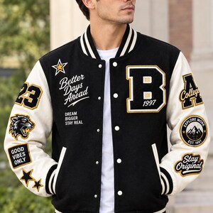 Peut inclure: Une veste universitaire noire et crème avec des boutons-pression blancs, des bords-côtes rayés et divers écussons brodés, dont la lettre B, le texte Better Days Ahead, Dream Bigger Stay Real, College et Good Vibes Only.