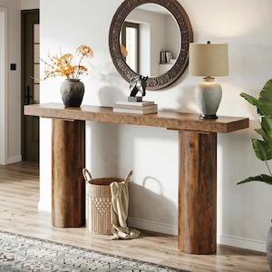 May include: A long wooden console table with a thick rectangular top supported by two large cylindrical wooden legs. The table holds a dark grey vase with orange flowers, a black horse sculpture on books, and a ribbed lamp with a beige shade.