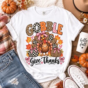 May include: A white t-shirt featuring a graphic of a turkey wearing a pink bow, surrounded by autumn leaves and two pumpkins. The text "GOBBLE" appears in a mix of leopard and checkered patterns, with "Give Thanks" written in black script below.