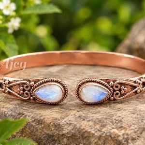 May include: A copper cuff bracelet featuring two teardrop-shaped moonstones at the open ends. The metal band displays intricate embossed scrollwork and beaded detailing surrounding each stone. The text AJCG is visible on the left side.