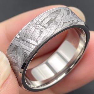 Puede incluir: Un anillo de metal en tono plateado con una banda exterior plana que presenta un patrón texturizado y grabado de líneas entrecruzadas y formas geométricas. El interior de la banda es liso, pulido y reflectante, con un acabado metálico.
