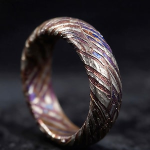May include: A ring featuring a textured, multi-layered surface with a pattern of swirling metallic colours including silver, copper, bronze, and deep purple. The band has a rugged, uneven finish with deep grooves and a high-contrast, layered appearance.