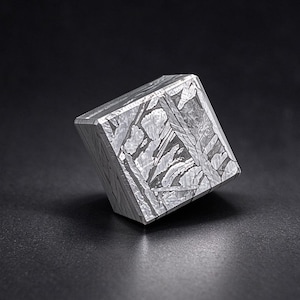 May include: A metallic cube featuring a distinct Widmanstätten pattern on its surface. The object displays a silver-grey color with intersecting crystalline lines and geometric etchings across its flat, polished faces.