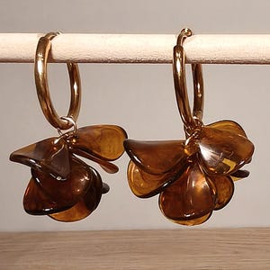 May include: A pair of gold-toned hoop earrings featuring dangling clusters of translucent, amber-colored, petal-shaped charms. The earrings have a polished metallic finish on the circular hoops and a smooth, glossy texture on the brown resin petals.