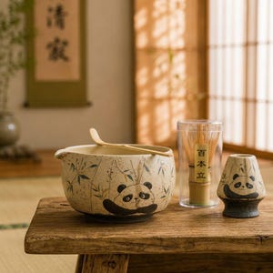 May include: A ceramic matcha bowl with a speckled cream finish, featuring a painted panda and bamboo stalks, accompanied by a bamboo whisk in a clear case, a bamboo scoop, and a matching ceramic panda whisk holder with a dark base.