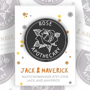 May include: A collection of round, black and gray enamel pins and matching cards. The pins and cards feature a rose design with the words "Rose Apothecary" and "Jack & Maverick" printed on them. The cards have a white background.