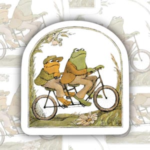 May include: A collection of stickers featuring a whimsical illustration of two frogs riding a tandem bicycle. The frogs are dressed in brown and tan clothing, with one wearing a hat. The background is a soft, neutral color with floral accents.