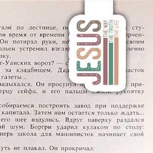 May include: A close-up of a hand holding a bookmark with the word "JESUS" in large, colorful letters. The bookmark is decorated with a colorful design and the words "The Way, The Truth, The Life".