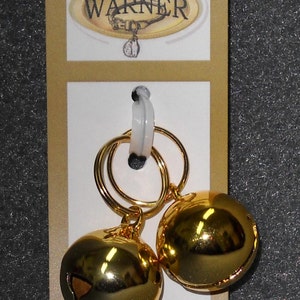 May include: Two gold-colored jingle bells with a white plastic tag attached. The tag has the word "WARNER" printed on it.