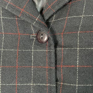 May include: A black blazer featuring a windowpane check pattern in thin red and white lines. The garment includes a notched lapel and a single dark brown button closure at the center front, showcasing a textured fabric weave throughout the piece.