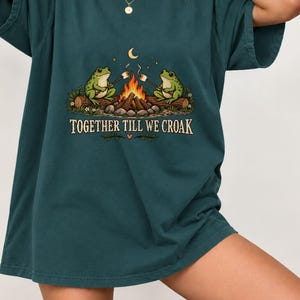 May include: A dark teal short-sleeve t-shirt featuring a graphic of two green frogs sitting on logs by a campfire roasting marshmallows. Above the frogs is a crescent moon and stars. The text TOGETHER TILL WE CROAK is printed below the fire.