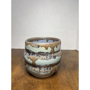 Peut inclure: Une tasse en céramique au corps arrondi et nervuré, dotée d'un émail mat bleu clair. L'extérieur est orné d'un motif de coulures couleur bronze métallique qui descend du bord et traverse les crêtes horizontales du récipient.