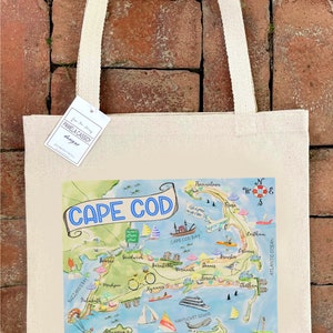 May include: Cream-colored tote bag featuring a colorful map of Cape Cod. The map includes illustrations of landmarks and towns, with the words "Cape Cod" in a blue banner. The bag has cream-colored handles and a tag.