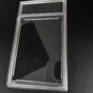 May include: A clear, rectangular plastic protective slab case designed for collectibles. The case features a thick, transparent frame with a dedicated rectangular window at the top for a label and a larger central window for displaying an item.
