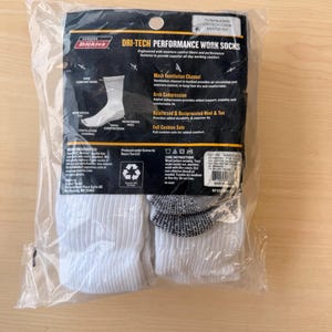 May include: A package of white Dickies Dri-Tech performance crew work socks in a clear plastic bag. The black cardboard header features product details including mesh ventilation channels, arch compression, and a reinforced heel and toe design.