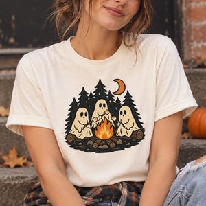 May include: A cream-colored short-sleeve t-shirt featuring a graphic print of three white ghosts sitting around a campfire in a forest. The design includes black pine trees, a small orange crescent moon, and the ghosts roasting marshmallows.
