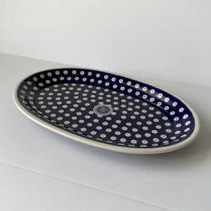 May include: An oval ceramic serving platter featuring a deep blue glaze decorated with a repeating pattern of small white dots with dark centers. A circular white sticker with text is centered on the interior surface of the dish.