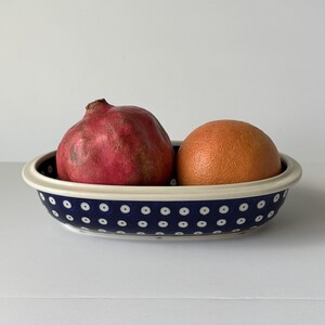 May include: An oval ceramic bowl with a deep blue exterior featuring a repeating pattern of small white dots. Inside the bowl sits one whole red pomegranate and one whole orange grapefruit. The bowl has a smooth, cream-colored rim.