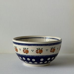 May include: A ceramic bowl featuring a cream-colored base with a hand-painted pattern of orange fruits with green leaves, accented by a dark blue band with white polka dots and a decorative border of small blue circles around the upper rim.