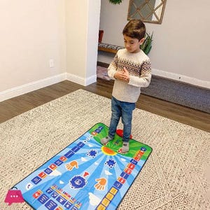 May include: A rectangular interactive prayer mat featuring a bright blue background with a yellow sun, white clouds, and orange hand icons. The mat includes various colorful squares with text and symbols arranged along the borders for educational use.