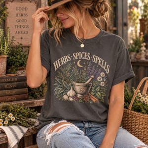 以下が含まれることがあります： 「HERBS SPICES SPELLS」という文字がプリントされたチャコールグレーのTシャツ。乳鉢と乳棒を中心に、様々なハーブ、花、ニンニク、シナモンスティックが描かれており、上部中央には小さな三日月のシンボルがあしらわれています。