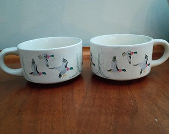May include: Two off-white ceramic mugs with a speckled finish, each featuring a repeating pattern of flying mallard ducks in shades of green, gray, and brown, accented by thin green reeds painted around the base of the exterior surface.