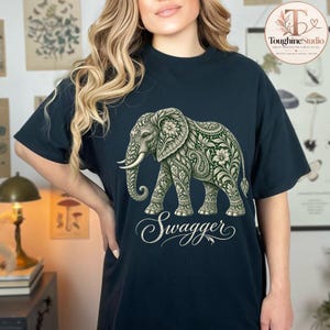 May include: A dark navy blue short-sleeve t-shirt featuring a large graphic of an elephant filled with intricate green and white floral and paisley patterns. Below the elephant, the word Swagger is printed in a white, elegant cursive script font.