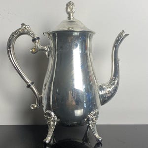 May include: A polished silver-plated coffee pot featuring an ornate handle with black accents, a curved spout with intricate floral detailing, a domed lid with a finial, and three decorative feet with embossed flower patterns at the base.