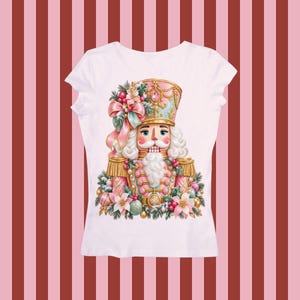 Puede incluir: Una camiseta blanca de manga corta con un gráfico central de un soldado cascanueces en tonos rosa pastel y dorado. El cascanueces está adornado con un lazo grande, ramas de pino, flores de pascua y adornos alrededor de su base.