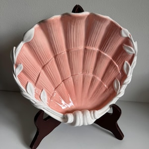 Puede incluir: Un cuenco de cerámica con forma de concha de vieira, con un interior rosa pastel brillante y un borde blanco con detalles en relieve en forma de hojas. El cuenco se exhibe sobre un soporte de plástico marrón oscuro, mostrando su textura estriada.