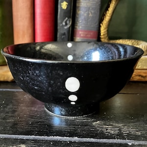 May include: A black ceramic bowl with a speckled finish and a vertical arrangement of three white circular dots of varying sizes on the exterior. The bowl features a wide, open rim and a circular base, presenting a smooth, glossy surface texture.