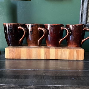 May include: A set of four ceramic mugs with a glossy, dark brown glaze and a slightly uneven, organic rim. Each mug features a curved handle and a tapered base, displaying variations in the brown tones across their smooth, reflective surfaces.