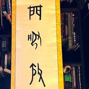 Puede incluir: Un pergamino de pared vertical que presenta un panel central de color crema con caracteres y símbolos caligráficos negros. El pergamino está enmarcado por un borde amarillo brillante con un patrón de dragón repetitivo y dos sellos rojos.