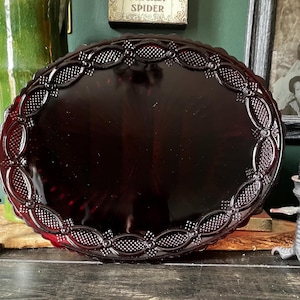 May include: An oval-shaped, deep red glass serving platter featuring a decorative pressed pattern along the rim. The border consists of repeating oval segments filled with a textured diamond grid design, separated by small circular accents.