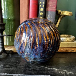 May include: A spherical ceramic object with a glossy finish featuring a swirling pattern of deep blue, metallic bronze, and dark brown glazes. The surface has a textured, rippled appearance with light reflecting off the curved, rounded form.