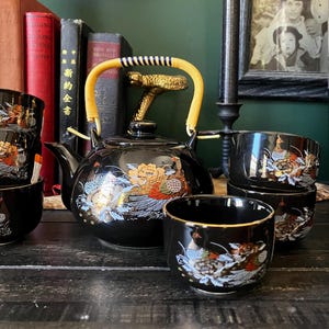 May include: A black ceramic tea set featuring a teapot with a yellow woven handle and matching tea cups. Each piece is decorated with a colorful pheasant and floral motif in gold, red, blue, and white tones, set against a glossy black finish.