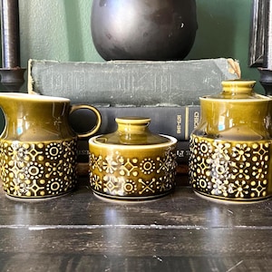 May include: A three-piece ceramic set featuring a creamer, a lidded sugar bowl, and a lidded canister, all finished in a glossy olive green glaze with a repeating white geometric star and circle pattern embossed around the lower sections.