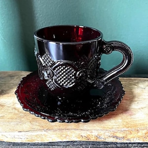 May include: A deep red glass teacup and matching saucer set featuring a prominent diamond-point hobnail pattern. The cup has a sturdy, curved handle with decorative detailing, and the saucer edge is scalloped with a textured floral motif.
