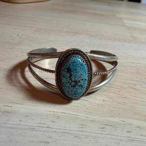 May include: A silver cuff bracelet featuring a central oval-shaped turquoise stone with a dark matrix pattern. The stone is framed by a twisted rope border, and the band consists of three separate silver wires that converge at the ends.