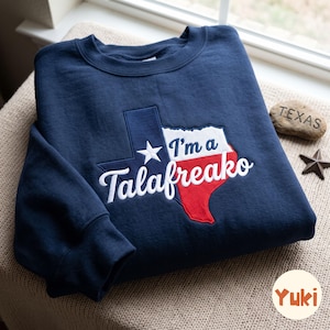 Pode incluir: Uma camisola de gola redonda azul-marinho com um gráfico bordado do estado do Texas em branco, azul e vermelho. O design inclui uma estrela branca e o texto "I'm a Talafreako" em letras cursivas brancas no centro da forma do estado.