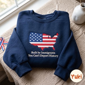 Può includere: Una felpa girocollo blu navy con una mappa ricamata degli Stati Uniti riempita con il motivo della bandiera americana. Sotto la mappa, il testo recita: Built by Immigrants You Can't Deport History in un carattere bianco senza grazie.