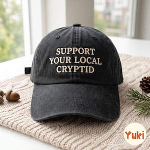 Puede incluir: Una gorra de béisbol de color gris carbón con el texto bordado "SUPPORT YOUR LOCAL CRYPTID" en color beige claro. La gorra cuenta con una corona estructurada, una visera curva y una correa de tela en la parte posterior ajustable.